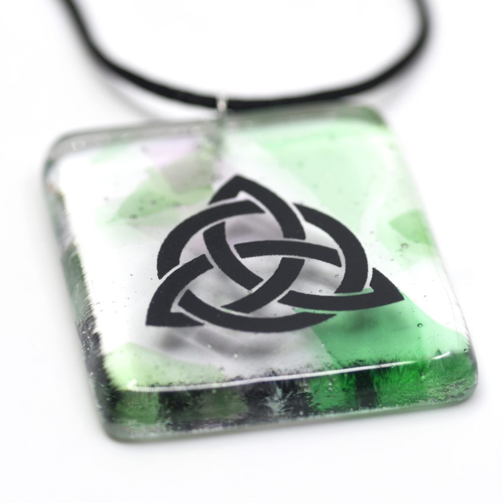 Fused glass Celtic Trinity Knot Pendant. The pendant is a clear glass with green and pink splashes of colour, with a detailed black Celtic knot.